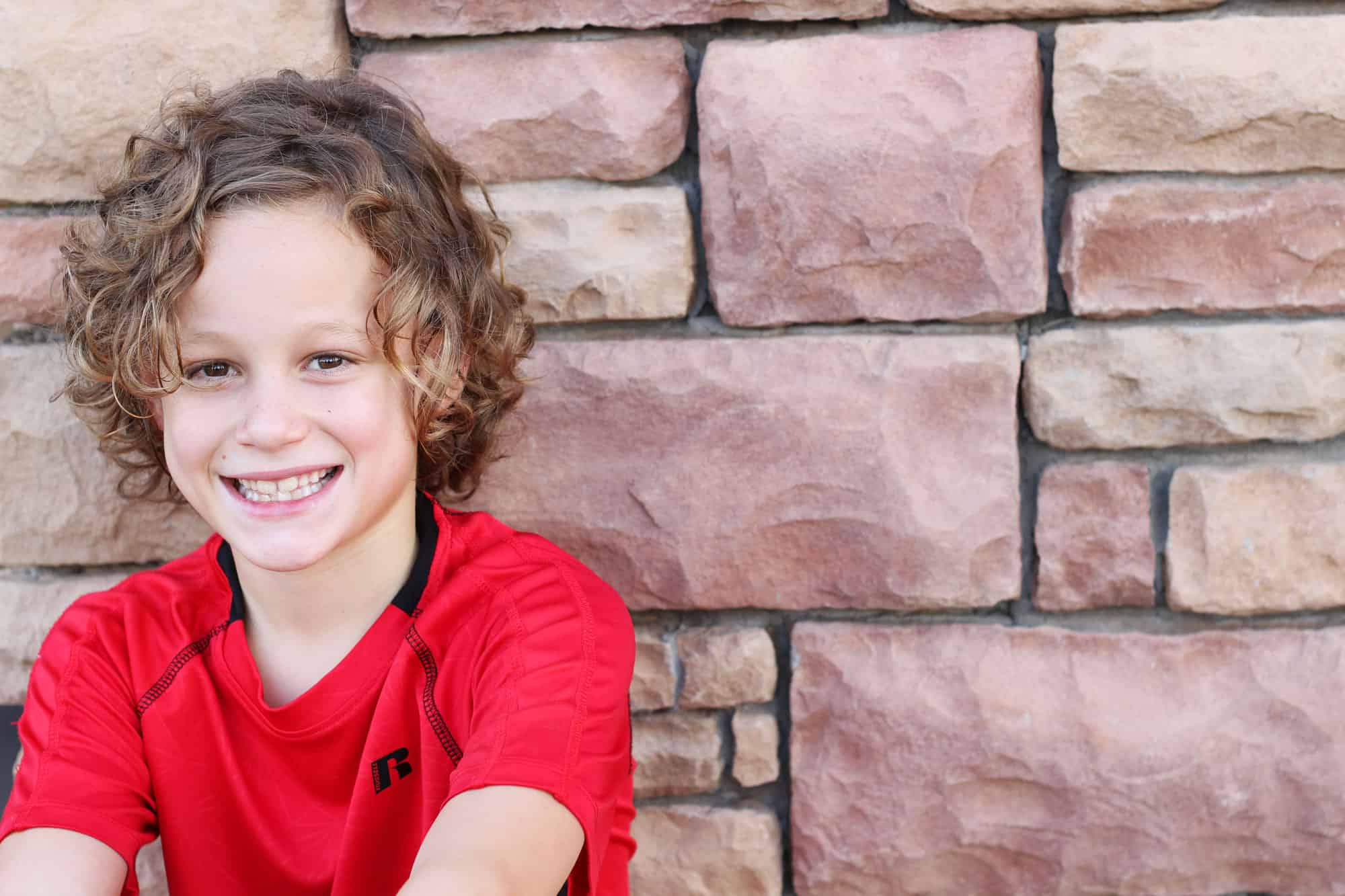 Boy in a red shirt sitting against a red brick wall.