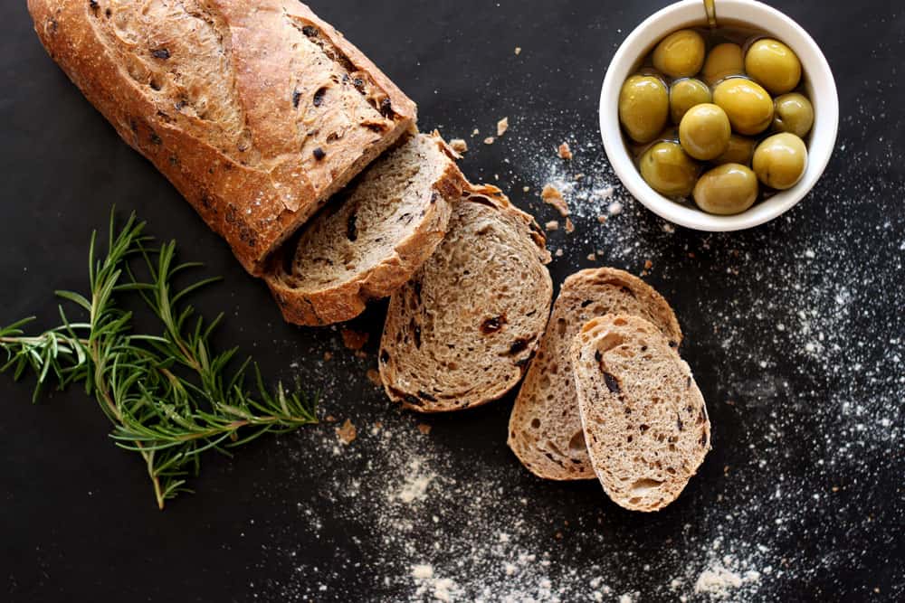 Loaf of olive bread sliced on a table, rosemary and green olives on table.