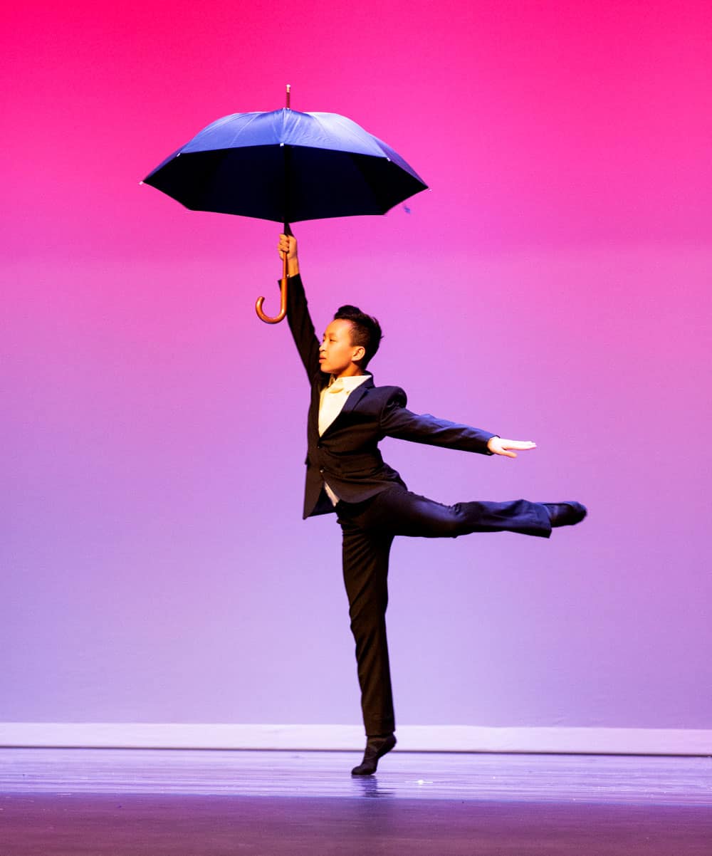 Solo dancer doing a back kick while holding a black umbrella while onstage.