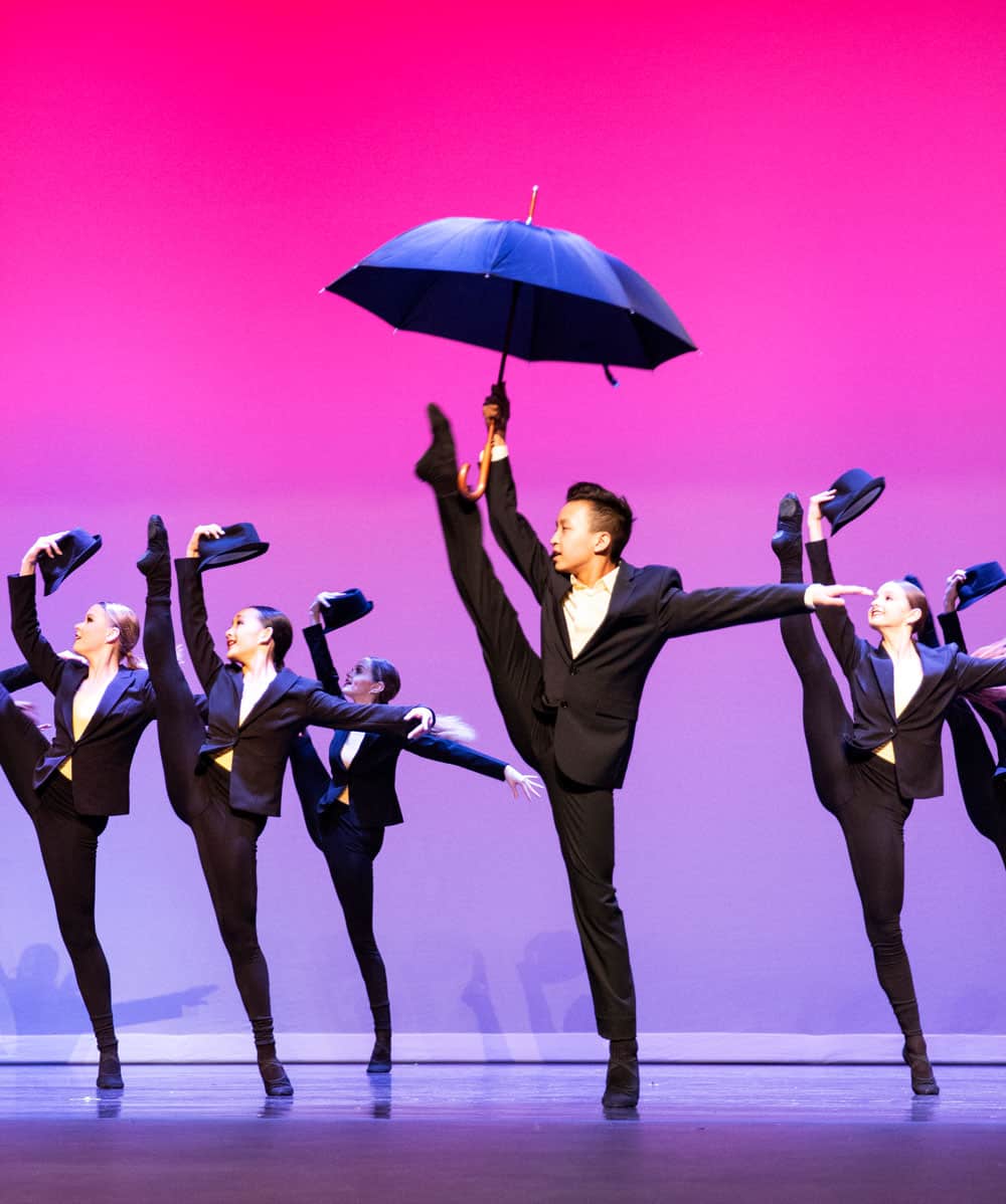 Musical theatre dance team performing on stage doing a leg kick, dancer holding an umbrella.